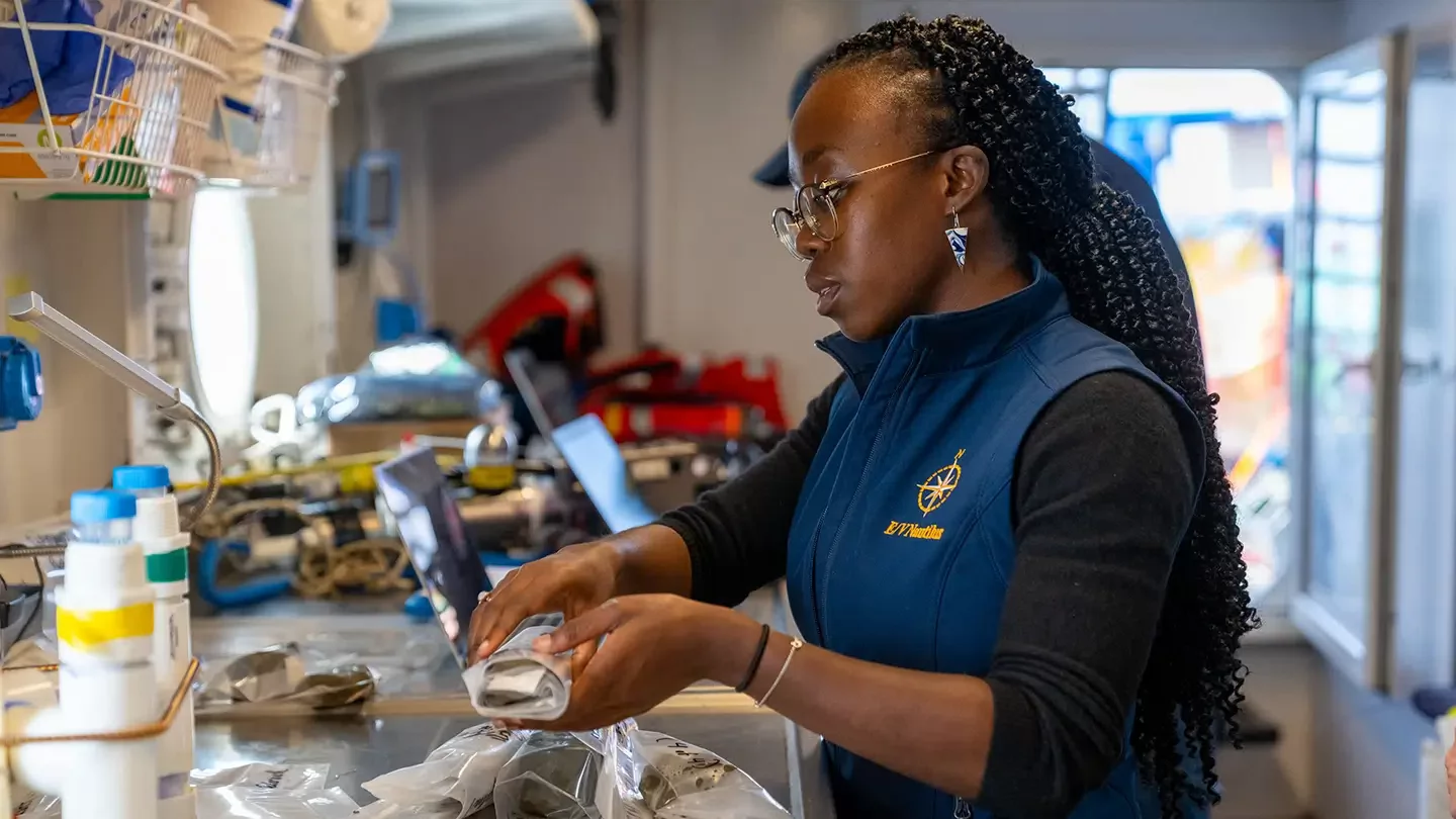 A woman with braided hair and glasses handles an object wrapped in plastic in a science lab.